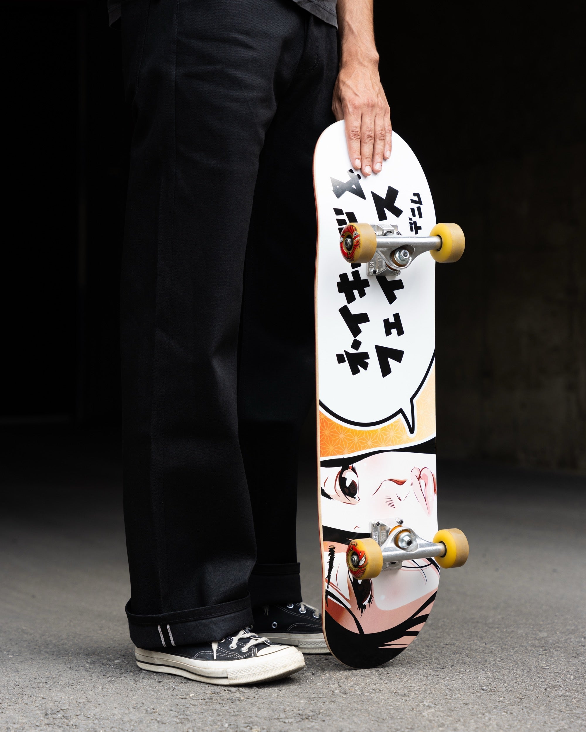 A person in black pants and black sneakers holds a skateboard with a white graphic deck featuring a speech bubble with Japanese text and an anime-style face illustration. The skateboard has yellow wheels and silver trucks, photographed against a dark concrete background.