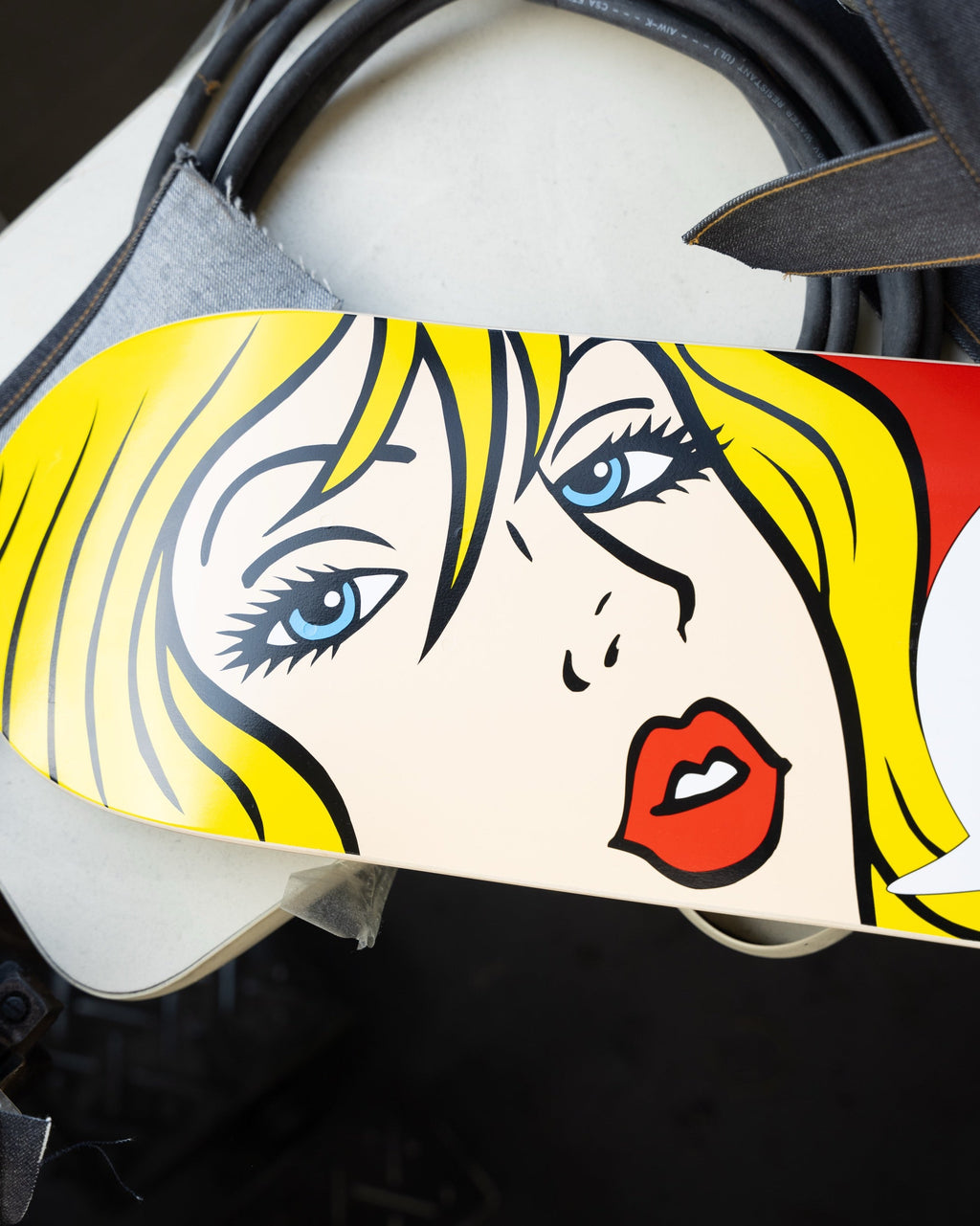 A close-up of a skateboard deck featuring a stylized pop-art illustration of a woman's face with yellow hair, blue eyes, black outlines, and red lips on a cream background.