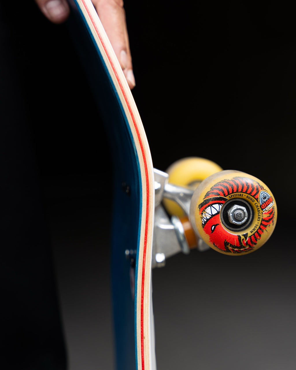Close-up of a skateboard deck edge with visible trucks and wheels, held by a hand against a dark background. The wheel has a red, yellow, and black graphic design.