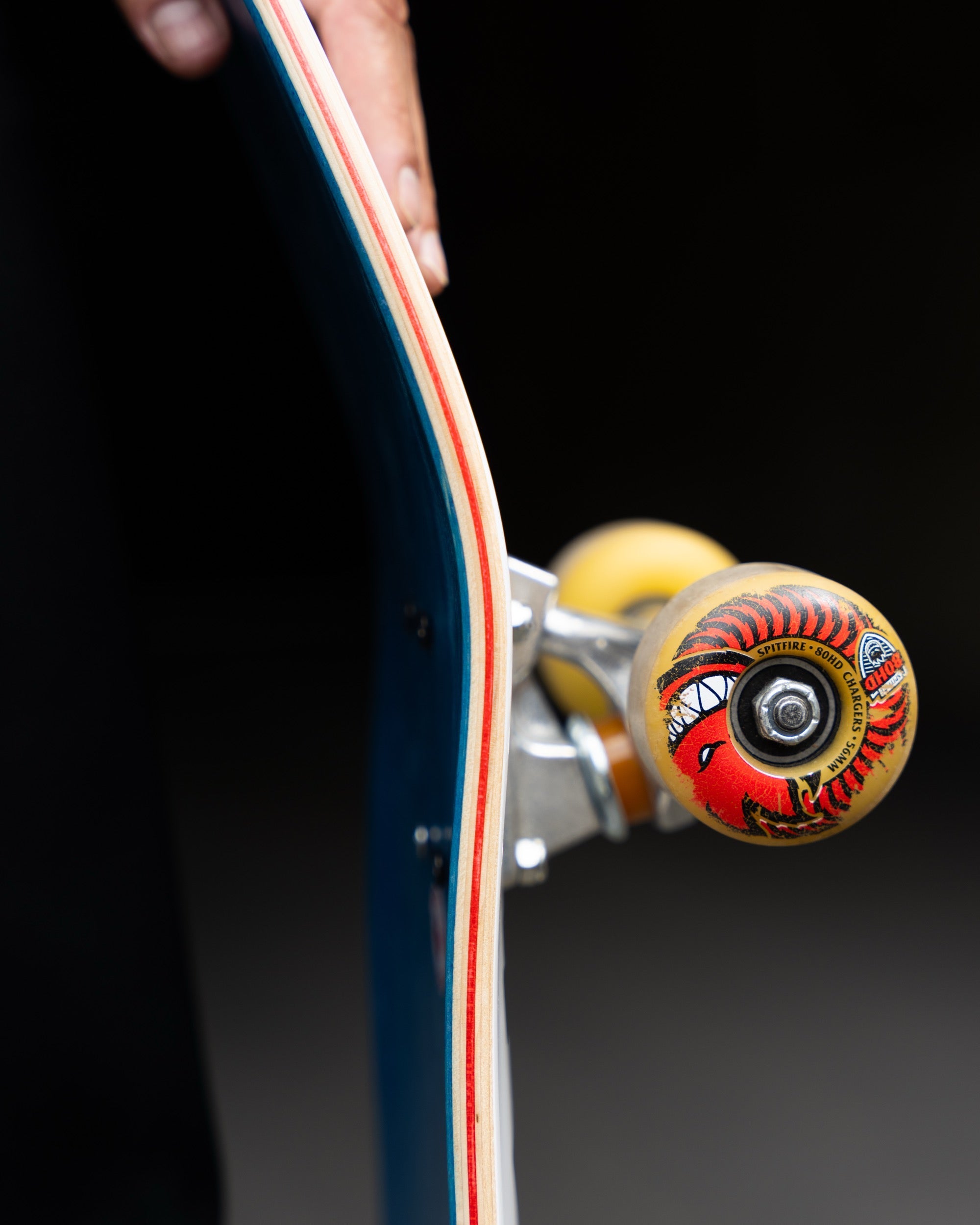 Close-up of a skateboard deck edge with visible trucks and wheels, held by a hand against a dark background. The wheel has a red, yellow, and black graphic design.