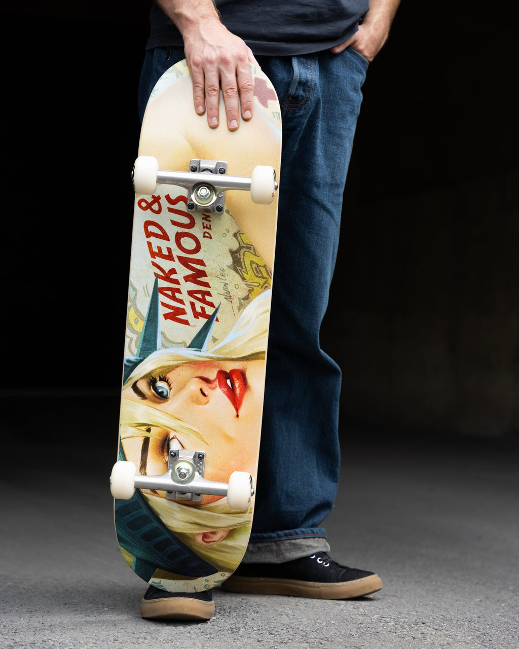 A person in blue jeans and black sneakers holds a skateboard with a vintage-style pin-up graphic featuring the text 'NAKED & FAMOUS DENMARK' on a dark studio background.